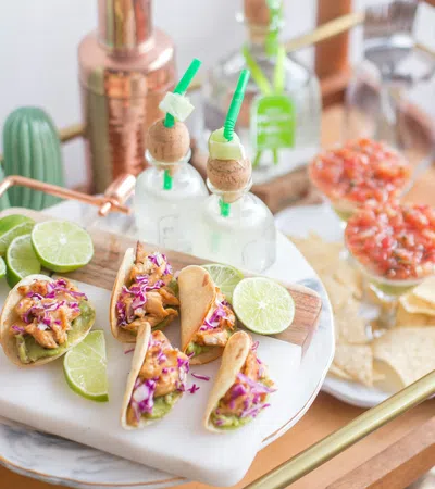 Mini chicken tacos with salsa, lime wedges, chips, and cocktails on a wooden and marble serving tray.