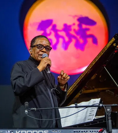 Jazz musician performing on stage with microphone and grand piano under colorful spotlight with dancing silhouettes.