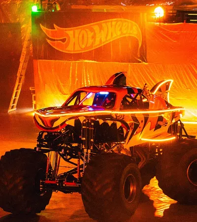 Monster truck with tiger stripes and glowing accents performs indoors in front of Hot Wheels banner at night.