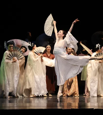 Ballet dancer in white costume performs with fans on stage with other dancers in traditional attire.
