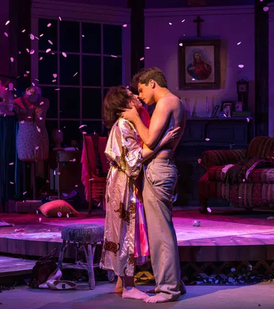 Couple embraces on stage in intimate theater scene with warm lighting and scattered petals.