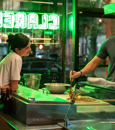 Man preparing pizza behind counter while woman watches inside a neon-lit pizzeria at night.