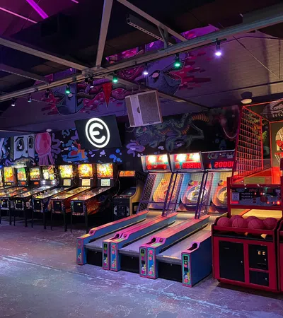 Indoor arcade with pinball machines, skee ball lanes, and basketball hoops under neon lighting.