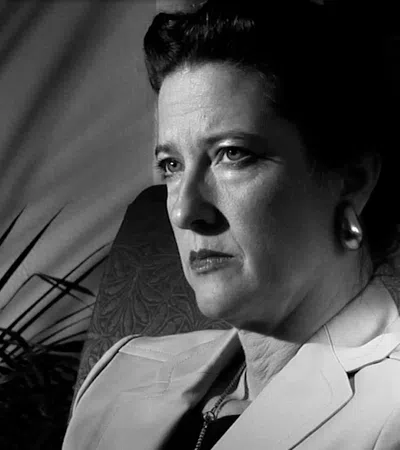 Black and white portrait of a serious woman sitting in a chair with shadows and a lamp in the background