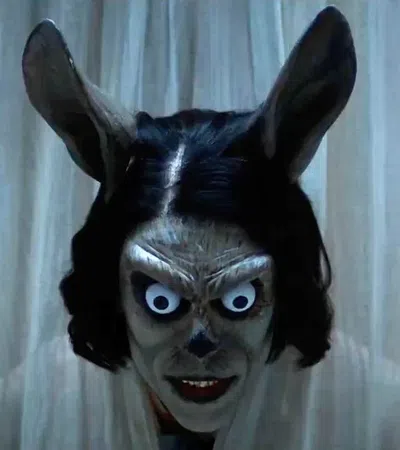 Creepy figure with exaggerated eyes, rabbit ears, and dark hair peers through white curtains in dim light.
