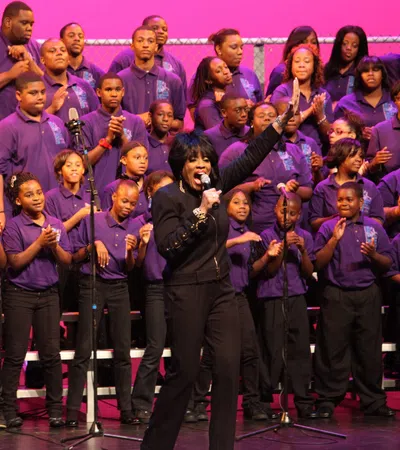 Choir of youths in purple shirts singing on stage with a soloist leading in black outfit and pink background.