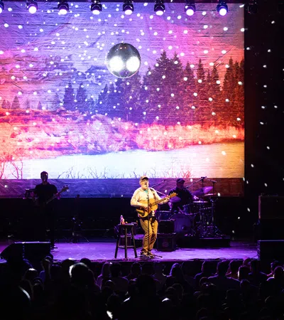 Band performing on stage with snow effects and forest landscape backdrop under disco ball lighting.