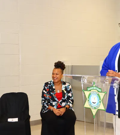 Man in blue cardigan speaking at podium with sheriff badge, woman in floral blazer seated nearby smiling