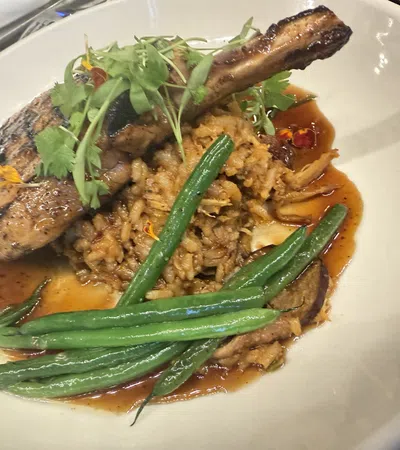 Grilled pork chop with green beans, rice, and sauce garnished with fresh herbs in a white bowl.
