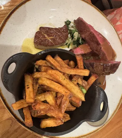Plate with grilled steak, sautéed greens, and a black dish filled with crispy golden French fries on a wooden table.