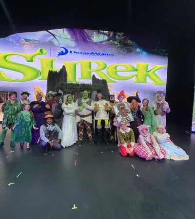 Cast of Shrek musical in full costumes posing on stage with Shrek show title backdrop