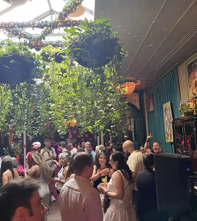 Crowded indoor party with hanging plants, colorful lighting, and guests socializing and enjoying drinks.