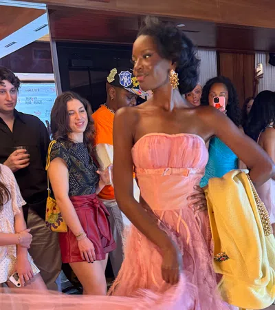 Model in pink gown holding yellow jacket walking past crowd of spectators at indoor event with wood paneling.