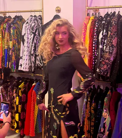 Woman in black dress posing in a colorful boutique with sequined and patterned jackets on racks