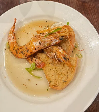 Two grilled shrimp served on toasted bread with broth and garnished with herbs on a white plate.