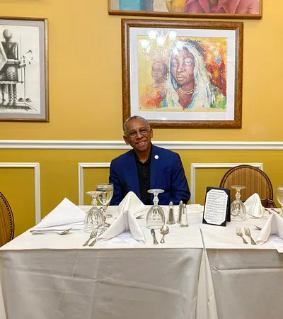 Man in blue suit sitting at white tablecloth-covered table set with wine glasses and silverware, framed art on yellow wall.