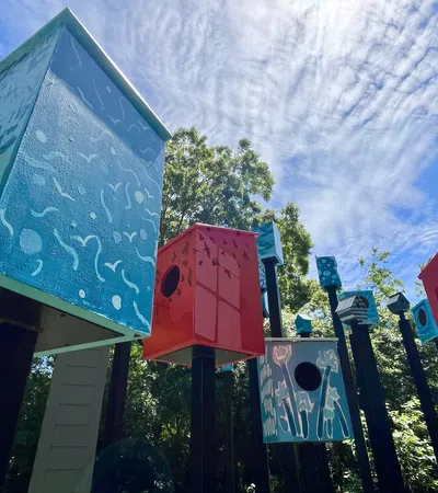 Colorful birdhouses mounted on black poles against a bright blue sky and leafy green trees.