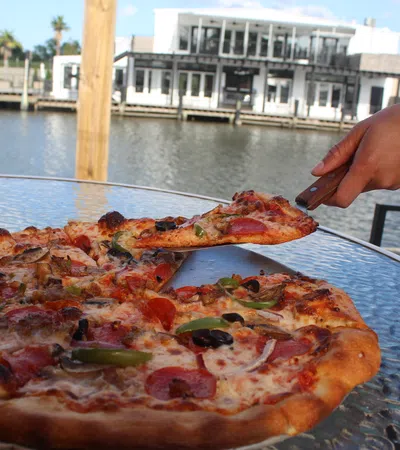 Hand picking a slice of pepperoni pizza with vegetables from a glass table by the waterfront.