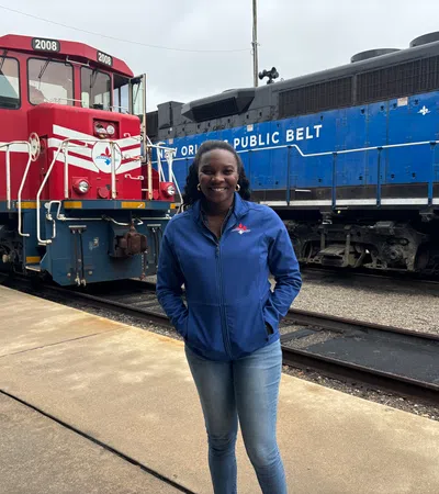 Smiling woman in blue jacket standing on platform between red and blue trains at a railway yard.