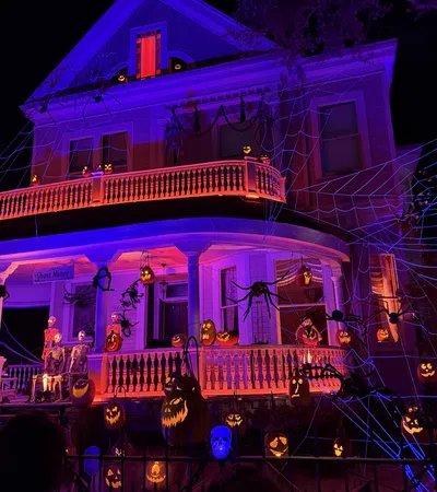 Large Victorian house decorated with glowing jack-o'-lanterns, skeletons, giant spiders, and web lights for Halloween night.