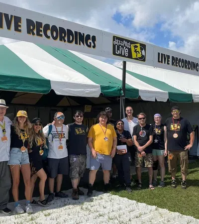 Group of people standing in front of a Jazz Fest Live tent offering live festival recordings during a sunny day.