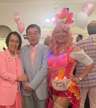 Two women in colorful circus costumes and pink wigs pose with a couple dressed in pastel pink and gray at a festive event.