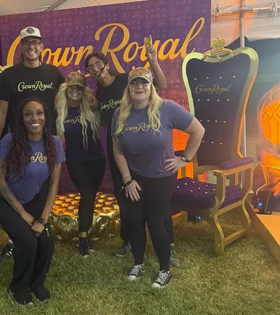 Five people wearing Crown Royal shirts pose in front of a Crown Royal backdrop between two large decorative royal chairs.