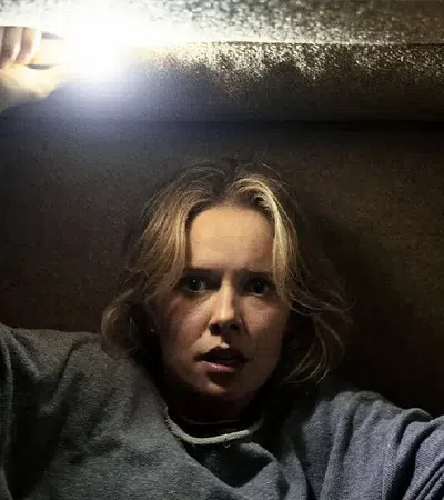 Woman trapped in a small dark space holding a bright flashlight above her head, looking fearful and tense.