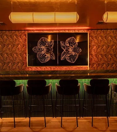 Dark bar interior with seven high chairs under warm lighting and orchid artwork on textured walls.