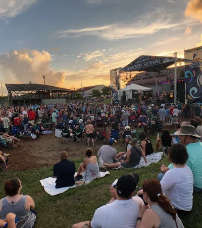Crowd enjoying live music at Jean Lafitte Seafood Festival during sunset with colorful sky and stage lighting.