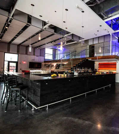 Modern industrial bar with black wooden counter, hanging pendant lights, glass railing staircase, and ambient lighting.
