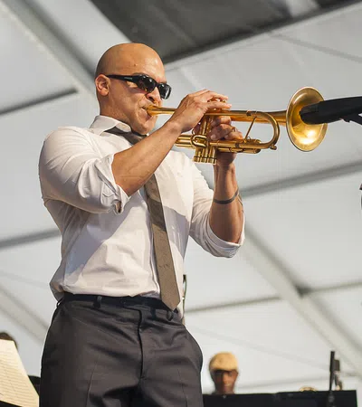 Bald man wearing sunglasses playing trumpet on stage under white tent with microphone nearby