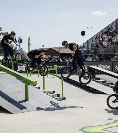 BMX rider performing a stunt sequence over a green rail in a skatepark with a large crowd watching.