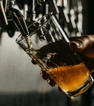 Close-up of a hand pouring amber beer into a tilted glass from a tap in a dimly lit bar.