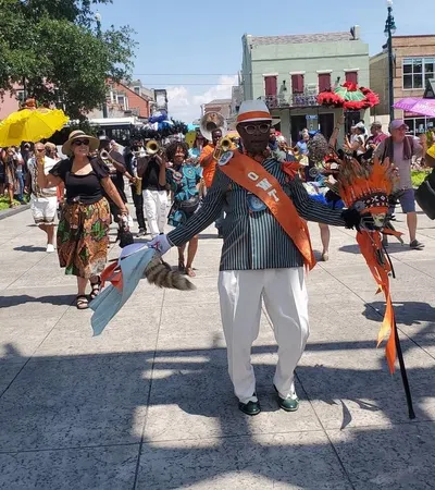 Man wearing a striped suit, orange sash, and hat leads a lively street parade with musicians and colorful umbrellas.