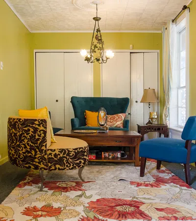 Cozy living room with patterned chairs, wooden fireplace, large floral rug, green walls, and natural light from windows.
