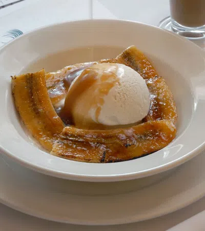 Grilled plantains topped with vanilla ice cream and caramel sauce in a white bowl on a white table.