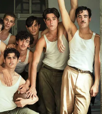 Group of eight young men wearing white tank tops and beige pants posing closely in a vintage room near open windows