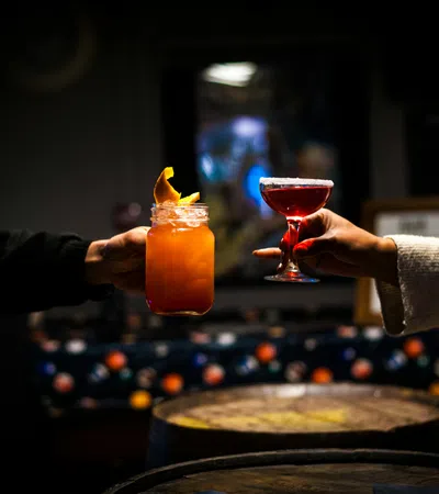 Two hands holding colorful cocktails in a dimly lit bar, clinking glasses over wooden barrels.