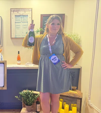 Woman holding wine bottle inside cozy wine bar with framed art, menu stand, plants, and candles in background.