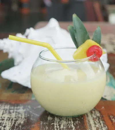 Pineapple smoothie in a round glass with a yellow straw, garnished with pineapple slice and cherry on a rustic wooden table.