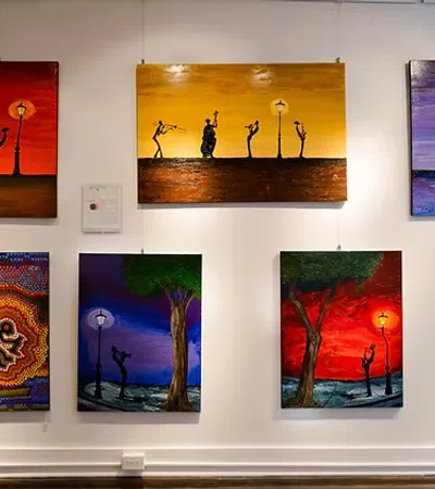 Colorful paintings of silhouetted musicians and dancers under street lamps displayed on a gallery wall.