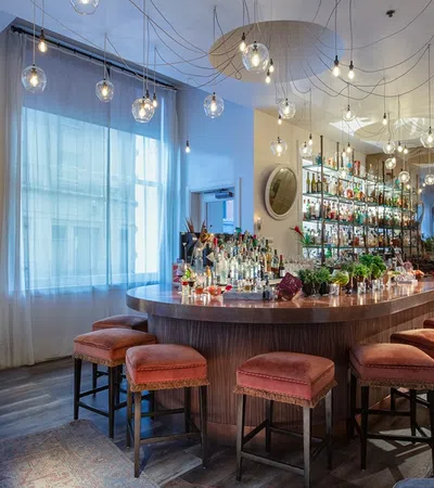 Modern upscale bar interior with curved wooden counter, cushioned stools, hanging lights, and stocked liquor shelves.