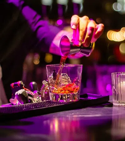 Bartender pours a red cocktail into a glass on a bar counter with dim purple lighting and bar tools nearby