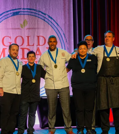 Group of chefs and officials posing with medals on stage at Gold Medal Plate culinary competition event.