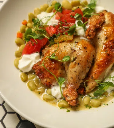 Roasted chicken leg quarters with white sauce, fava beans, diced tomatoes, and microgreens on white plate.