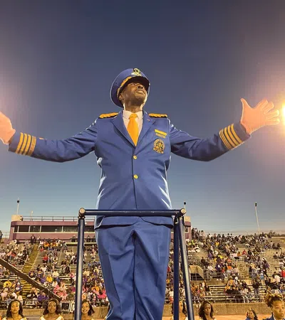 Band director in blue uniform conducting with baton at a stadium filled with spectators during evening.