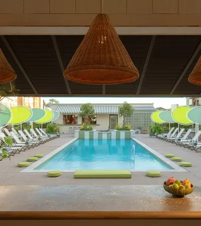 Outdoor pool area with lounge chairs, green sunshades, potted plants, and woven pendant lights over a bar counter.