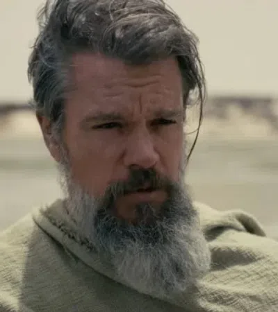 Man with gray beard and hair wearing a green cloak in a desert landscape with blurred sandy background.
