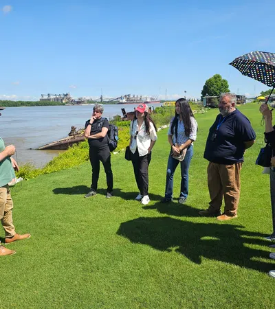 Group of six adults listening to a man speaking by a riverside on a sunny day with clear blue sky.
